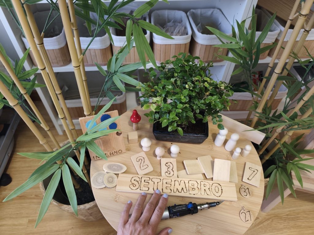 Mão a trabalhar numa composição artesanal em madeira, rodeada de plantas e materiais do ateliê AKSHARA.